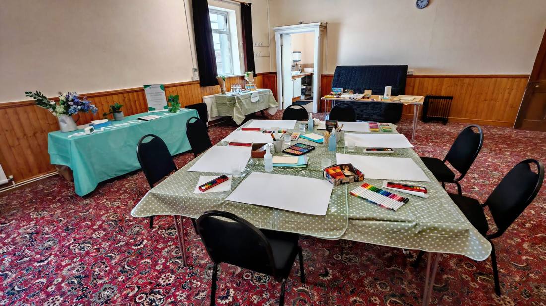A community hall set up for an art session, with paper, pastels and paints laid out on a polka-dot tablecloth